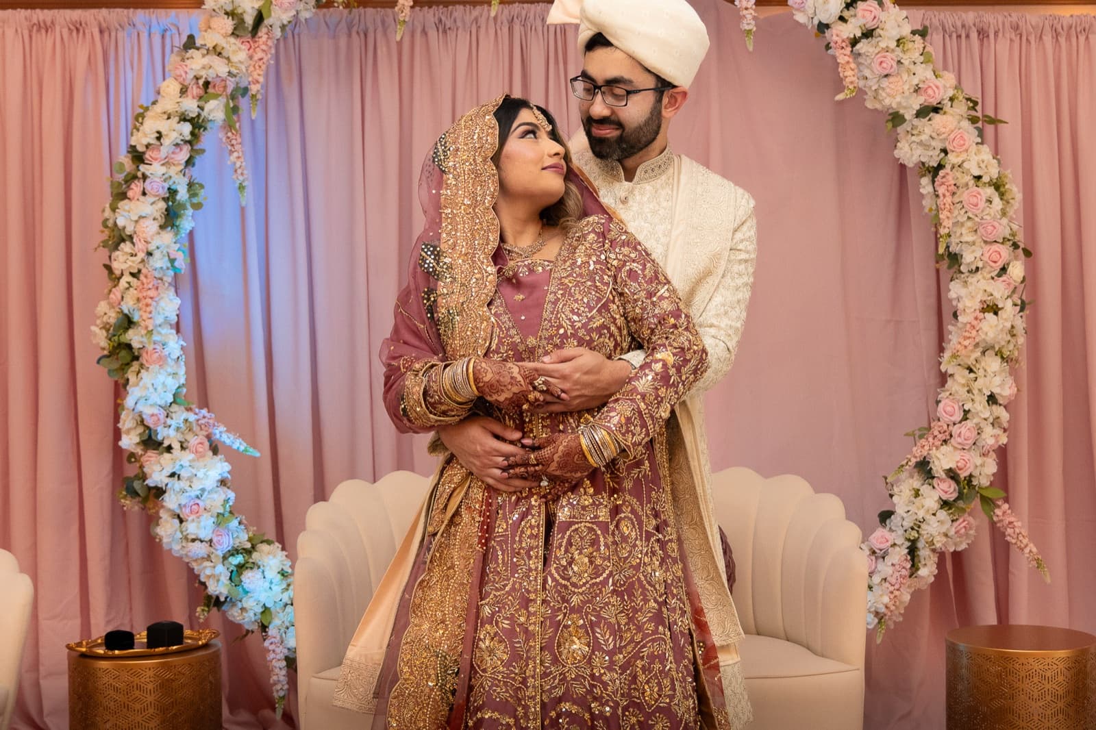 Faisal wrapping Ayesha from behind under the floral hoop, her henna hands over his